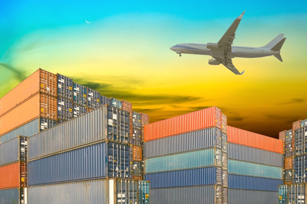 Photo of an aeroplane flying over stacked shipping containers.