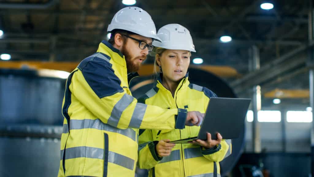 Two colleagues wearing hard hats and hi-vis discuss factoring and invoice discounting with a laptop in a large warehouse space
